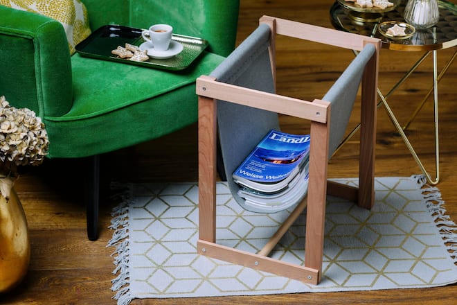 Zeitungsständer aus Holz mit Zeitschriften neben einem Sessel, auf dem ein Tablett mit einer gefüllten Kaffeetasse und Keksen steht